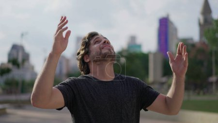 Foto de Feliz hombre levantando los brazos al cielo agradeciendo a Dios con ESPERANZA y FE. Un hombre caucásico de unos 30 años sintiendo GRATITUD parado al aire libre mirando hacia el parque - Imagen libre de derechos