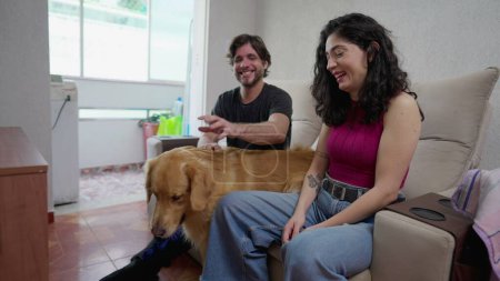 Foto de Auténticos dueños de perros felices riendo y sonriendo mientras están sentados en el sofá en interiores en una relación amorosa con la mascota Golden Retriever. Mujer juega con pelota y perro - Imagen libre de derechos