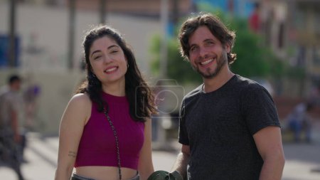 Téléchargez les photos : Joyeux couple debout dehors dans la rue de la ville souriant à la caméra au soleil. Millénaire homme et femme dans les années 30 posant sentiment heureux - en image libre de droit