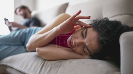 Foto de Mujer preocupada acostada en el sofá con una expresión fatigada mientras su novio mira al teléfono en segundo plano. Persona femenina en sufrimiento mental - Imagen libre de derechos