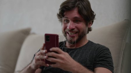 Photo for Happy man reacting positively to news notification on cellphone device while sitting at home couch. Ecstatic person celebrating success - Royalty Free Image