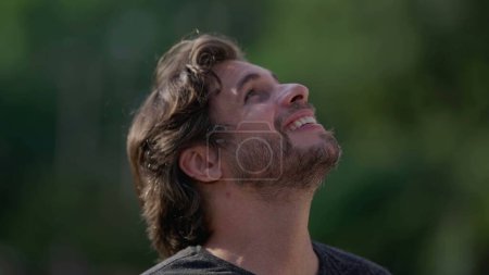 Téléchargez les photos : Un jeune homme heureux levant les yeux vers le ciel avec espoir et foi. Homme souriant regarde vers le haut avec GRATITUDE à l'extérieur dans le suivi plan rapproché visage - en image libre de droit