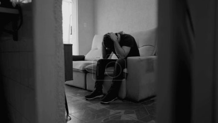 Photo for Lonely young man sitting on sofa inside apartment feeling alone. Anxious male person in 30s suffering from depression in dramatic monochromatic black and white - Royalty Free Image