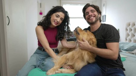 Foto de Pareja jugando con Dog en casa sentada en la cama. Joven hombre y mujer interactuando con su mascota Golden Retriever - Imagen libre de derechos