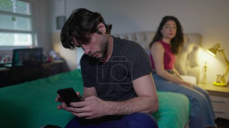 Foto de Pareja desconectada. Novio mirando el teléfono ignorando a su novia en el dormitorio. Hombre no comunicativo absorbido por las redes sociales en línea, mientras que la mujer quiere atención - Imagen libre de derechos