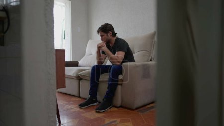 Téléchargez les photos : Jeune homme inquiet assis sur le canapé à la maison appartement avec expression préoccupée coûteuse. Concept de solitude de la personne masculine solitaire à l'intérieur - en image libre de droit