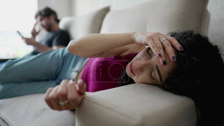 Téléchargez les photos : Jeune femme inquiète couchée sur un canapé souffrant de douleur émotionnelle. Femme en détresse dans les années 30 se sentant déconnecté de son petit ami en arrière-plan regardant son téléphone - en image libre de droit