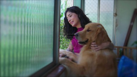 Téléchargez les photos : Femme heureuse dans la trentaine avec son chien regardant par la fenêtre de l'appartement. Une femme regardant dehors avec son Golden Retriever - en image libre de droit