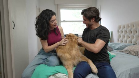 Photo for Happy couple interacting with their Golden Retriever Pet indoors. Man and woman petting their Dog while sitting in bedside at apartment - Royalty Free Image