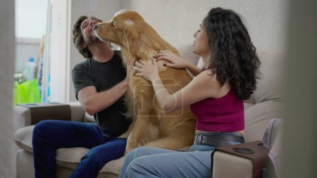 Téléchargez les photos : Drôle de moment d'amour de chien léchant propriétaire masculin dans le visage. Candid Couple assis sur le canapé à la maison avec Golden Retriever - en image libre de droit