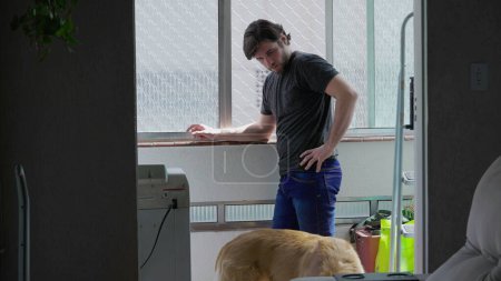 Foto de Un joven pensativo y reflexivo de pie junto a la ventana dentro de su apartamento. La vida doméstica de un hombre de 30 años mirando desde el balcón - Imagen libre de derechos