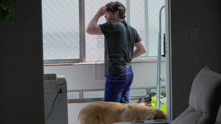 Téléchargez les photos : Un jeune homme cher regardant par la fenêtre à l'intérieur de l'appartement salon. Style de vie domestique franc et authentique - en image libre de droit