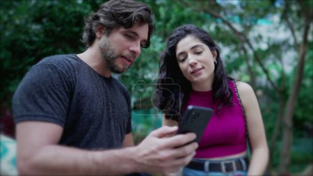 Photo for Young man showing online content to woman on cellphone device, both standing outside at city park. Two people looking at smartphone screen comenting on the media they are watching - Royalty Free Image