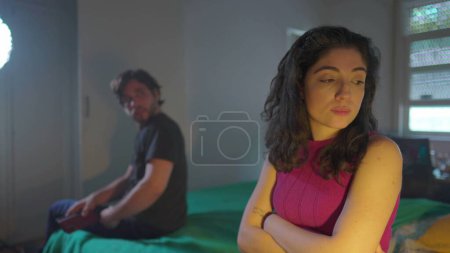 Foto de Mujer molesta cruzando los brazos ignorando a su pareja masculina sentada al lado de la cama en el dormitorio. Novia no comunicativa despedida novio - Imagen libre de derechos