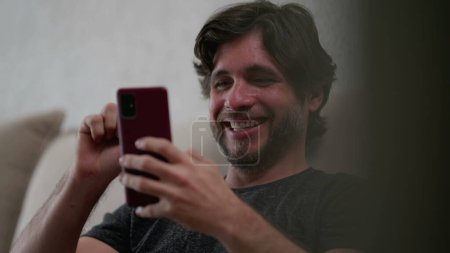 Téléchargez les photos : Homme heureux regardant l'appareil smartphone à la maison. Homme dans la trentaine assis sur le canapé défilant à travers les médias sociaux avec téléphone portable à la main - en image libre de droit