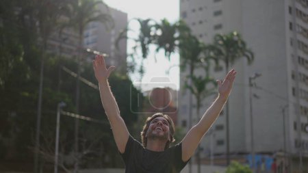 Téléchargez les photos : Un jeune homme heureux contemplatif acclamant le succès tout en se tenant debout à l'extérieur levant les bras dans l'air sentiment de bonheur et de gratitude - en image libre de droit