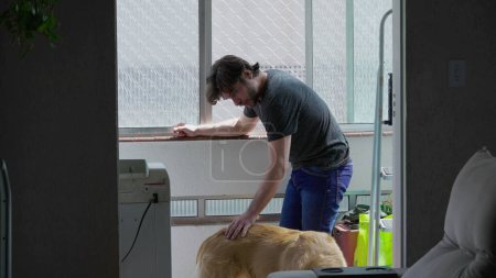 Téléchargez les photos : Jeune homme caressant son chien à la maison appartement par fenêtre. Candide homme avec son Golden Retriever Pet à l'intérieur - en image libre de droit