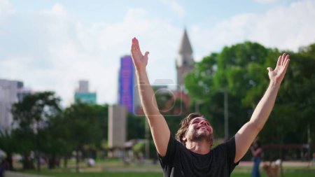 Foto de Joven fiel levanta los brazos al cielo sintiéndose GRATUITO al aire libre en la ciudad urbana. Un hombre celebra la VIDA con ESPERANZA - Imagen libre de derechos