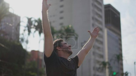Photo for One contemplative happy young man cheering success while standing outdoors raising arms in the air feeling happiness and gratitude - Royalty Free Image