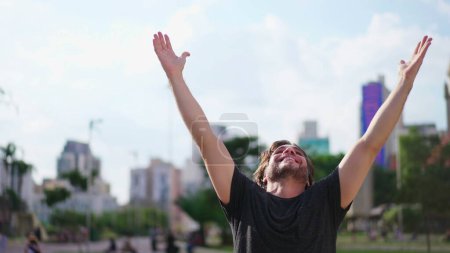Foto de Joven fiel levanta los brazos al cielo sintiéndose GRATUITO al aire libre en la ciudad urbana. Un hombre celebra la VIDA con ESPERANZA - Imagen libre de derechos
