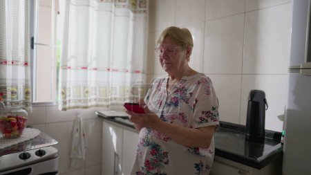 Téléchargez les photos : Femme âgée utilisant un téléphone portable tout en s'appuyant sur un évier de cuisine. Vieillesse domestique scène de style de vie de la personne âgée dans les années 80 navigation sur Internet en ligne - en image libre de droit