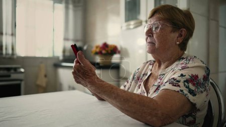 Foto de Anciana mujer caucásica involucrada en el estilo de vida doméstico, desplazándose a través de las redes sociales en el teléfono en la cocina casera - Imagen libre de derechos
