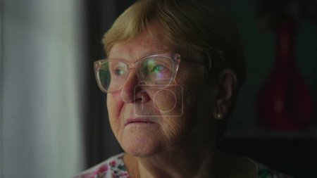 Téléchargez les photos : Femme âgée retraitée regardant par la fenêtre dans une profonde réflexion mentale. Gros plan sur le visage d'une femme contemplative dans les années 80 - en image libre de droit