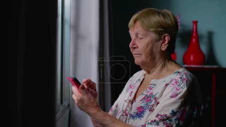 Téléchargez les photos : Femme âgée utilisant un téléphone intelligent par fenêtre à la maison, Message de dactylographie d'une personne âgée, Intersection de la technologie moderne et de la vieillesse - en image libre de droit