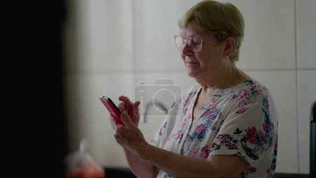 Téléchargez les photos : Femme âgée naviguant sur Internet sur téléphone cellulaire, Tech-Savvy Senior engagé avec la technologie moderne à la maison - en image libre de droit