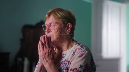 Foto de Mujer mayor religiosa esperanzada orando a Dios en el dormitorio, sintiéndose conectada a un poder superior y devota en oración - Imagen libre de derechos