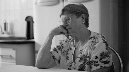 Téléchargez les photos : Femme âgée déprimée dans les années 80 exprimant solitude et solitude, assise à la cuisine en noir et blanc monochrome - en image libre de droit