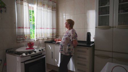 Téléchargez les photos : Contemplant femme âgée debout près de la fenêtre de la cuisine avec une expression réfléchie. Charmant style de vie domestique authentique vieillesse, rêveur des années 80 - en image libre de droit