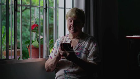 Téléchargez les photos : Femme âgée franche utilisant un appareil smartphone par fenêtre à la maison, éclairage spectaculaire de la personne âgée dans les années 80 en tapant un message sur le téléphone, la technologie moderne et la vieillesse - en image libre de droit