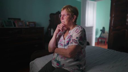Téléchargez les photos : Femme âgée à la retraite réfléchissant à la solution au problème tout en étant assis dans la chambre à coucher à côté du lit dans la solitude et la contemplation, vie de vieillesse authentique domestique - en image libre de droit