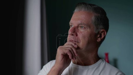 Foto de Hombre de mediana edad con el pelo gris mirando por la ventana con la mano en la barbilla, expresión contemplativa que sugiere un pensamiento profundo sobre la vida - Imagen libre de derechos
