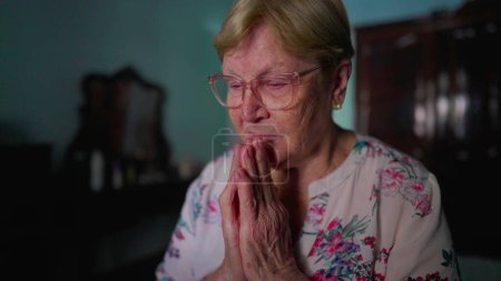 Téléchargez les photos : Femme âgée pleine d'espoir priant Dieu, assise près du lit dans la chambre à coucher, style de vie domestique authentique reflétant la vieillesse et la foi - en image libre de droit