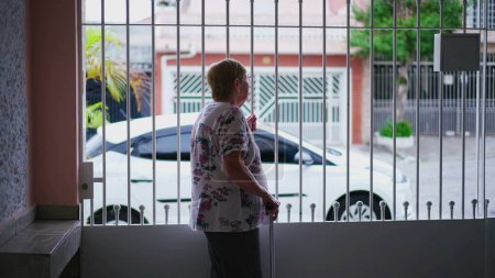 Téléchargez les photos : Observation de la maison de la femme âgée debout à l'intérieur de la maison fermée, surplombant le trottoir, s'accrochant à la barre métallique, regardant la montre de quartier - en image libre de droit