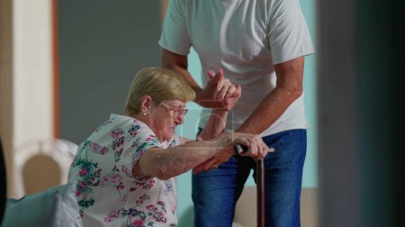 Photo for Person assisting elderly woman to get up from couch and walking through home corridor with the help of walking stick grip. Authentic depiction of old age lifestyle - Royalty Free Image