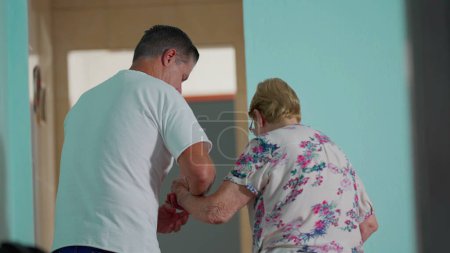 Photo for Person assisting elderly woman to get up from couch and walking through home corridor with the help of walking stick grip. Authentic depiction of old age lifestyle - Royalty Free Image