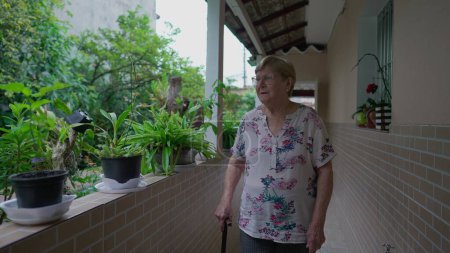Téléchargez les photos : Femme blanche âgée réfléchie dans les années 80 marchant à la maison, contemplant le jardin avec un bâton de marche - en image libre de droit