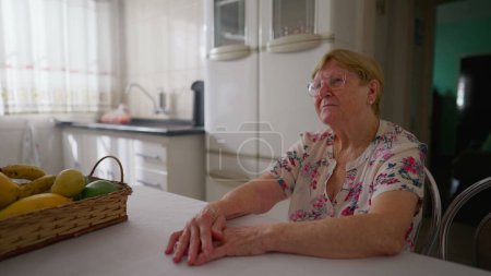 Photo for Contemplative elderly woman sitting at home kitchen gazing with pensive expression. Authentic real life domestic scene lifestyle of an aged caucasian 80s senior lady - Royalty Free Image