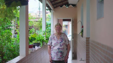 Téléchargez les photos : Casual Senior femme se tient à la maison arrière-cour en regardant la caméra. Suivi plan d'une personne âgée caucasienne âgée dans son 80S - en image libre de droit