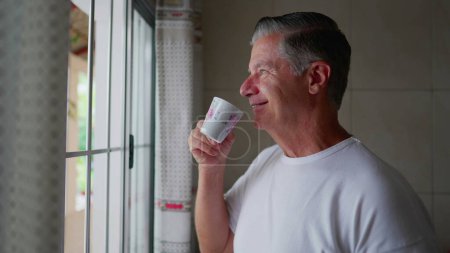Foto de Contemplativo hombre de mediana edad de pie junto a la ventana de la cocina bebiendo café durante el ritual matutino, estilo de vida diario doméstico de gris hombre de pelo sorbe bebida caliente - Imagen libre de derechos