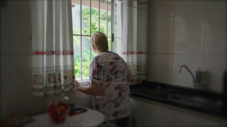 Foto de Mujer mayor abriendo cortinas de pie en la cocina. Ritual matutino de la mujer mayor que comienza la rutina del día y mira hacia fuera a la vista - Imagen libre de derechos