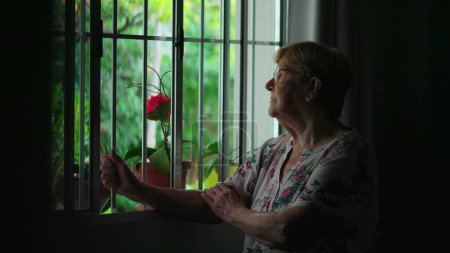 Foto de Mujer mayor con expresión pensativa sosteniendo la barra de la ventana, escena contemplativa del estilo de vida doméstico auténtico de la vejez - Imagen libre de derechos