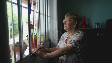 Téléchargez les photos : Femme âgée avec l'expression pensive tenant la barre de fenêtre, scène contemplative de la vieillesse authentique style de vie domestique - en image libre de droit