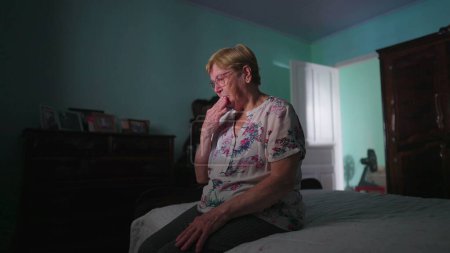 Foto de Religiosa anciana haciendo la señal de la cruz mientras sititng al lado de la cama en el dormitorio y orando a DIOS. Persona esperanzada y fiel en la vejez - Imagen libre de derechos