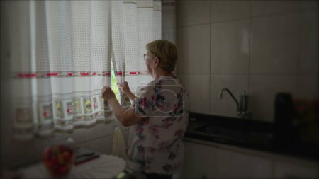 Téléchargez les photos : Femme âgée ouvrant rideaux debout à la cuisine. Rituel matinal de la femme âgée commençant la routine de la journée et regardant à la vue - en image libre de droit