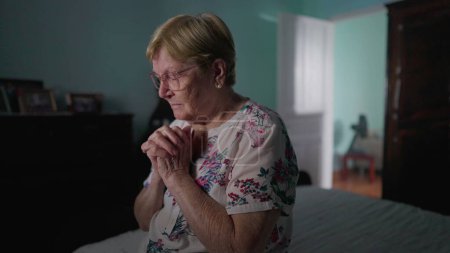 Foto de Anciana religiosa sentada junto a la cama en oración. HOPEFUL señora mayor Orando a DIOS en el dormitorio, estilo de vida doméstico auténtica vida real que representa la vejez en FE - Imagen libre de derechos