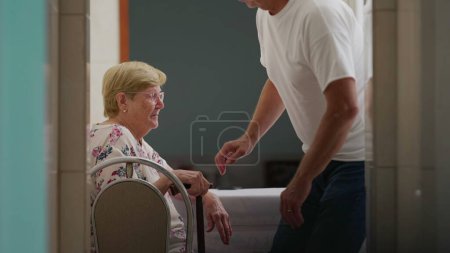 Photo for Assisting Elderly woman at home, candid authentic scene of adult son helping senior mother to get up from chair and walk through corridor - Royalty Free Image